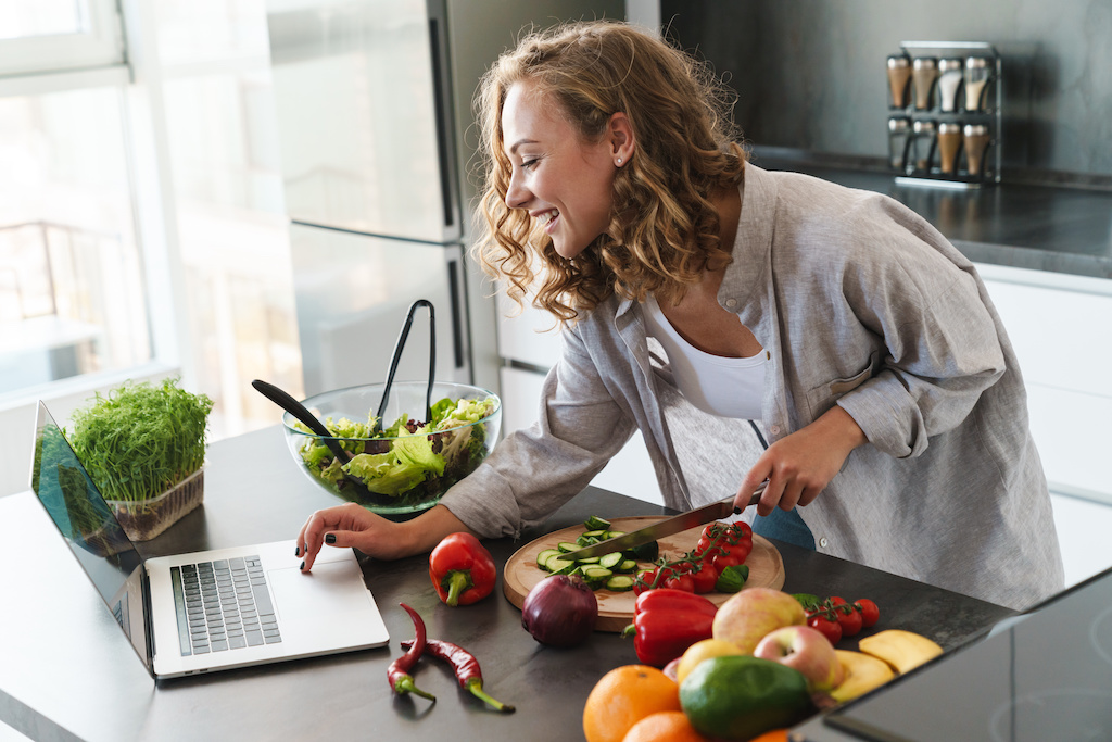 Happy young woman making a salad at the kitchen, chopping vegetables, looking at laptop computer