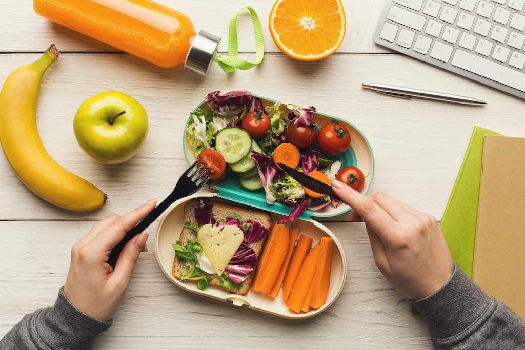 Healthy snack at office workplace. Businesswoman eating organic vegan meals from take away lunch box at wooden working table with computer keyboard